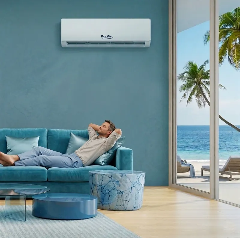 Man reclining on a teal sofa in a seaside Accra apartment with a PUR AIR split unit on the wall and palms framing the ocean outside.
